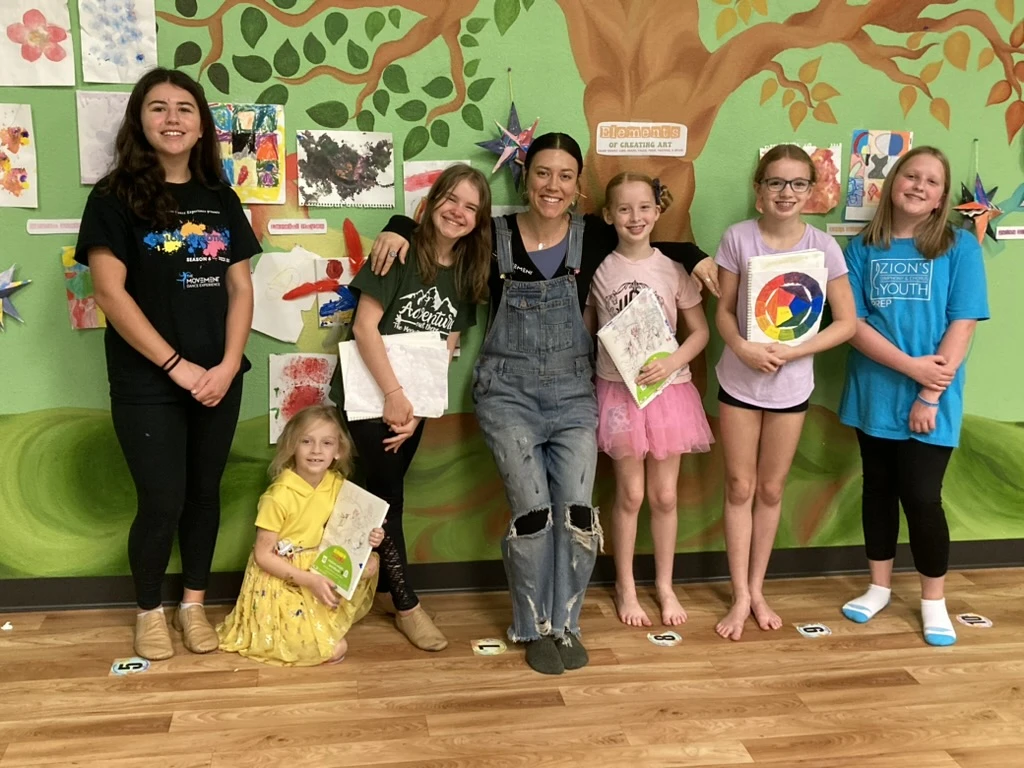 ArtHaus artists proudly holding their sketchbooks with their teacher in front of the student art wall