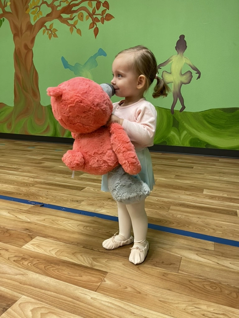 A toddler in ballet slippers hugging a big pink teddy bear