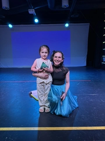 A little dancer proudly holding her award with her teacher on the Ovation Theatre stage